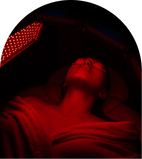 Person lying under a red light therapy device, eyes shielded, wrapped in a towel, in a dimly lit setting.