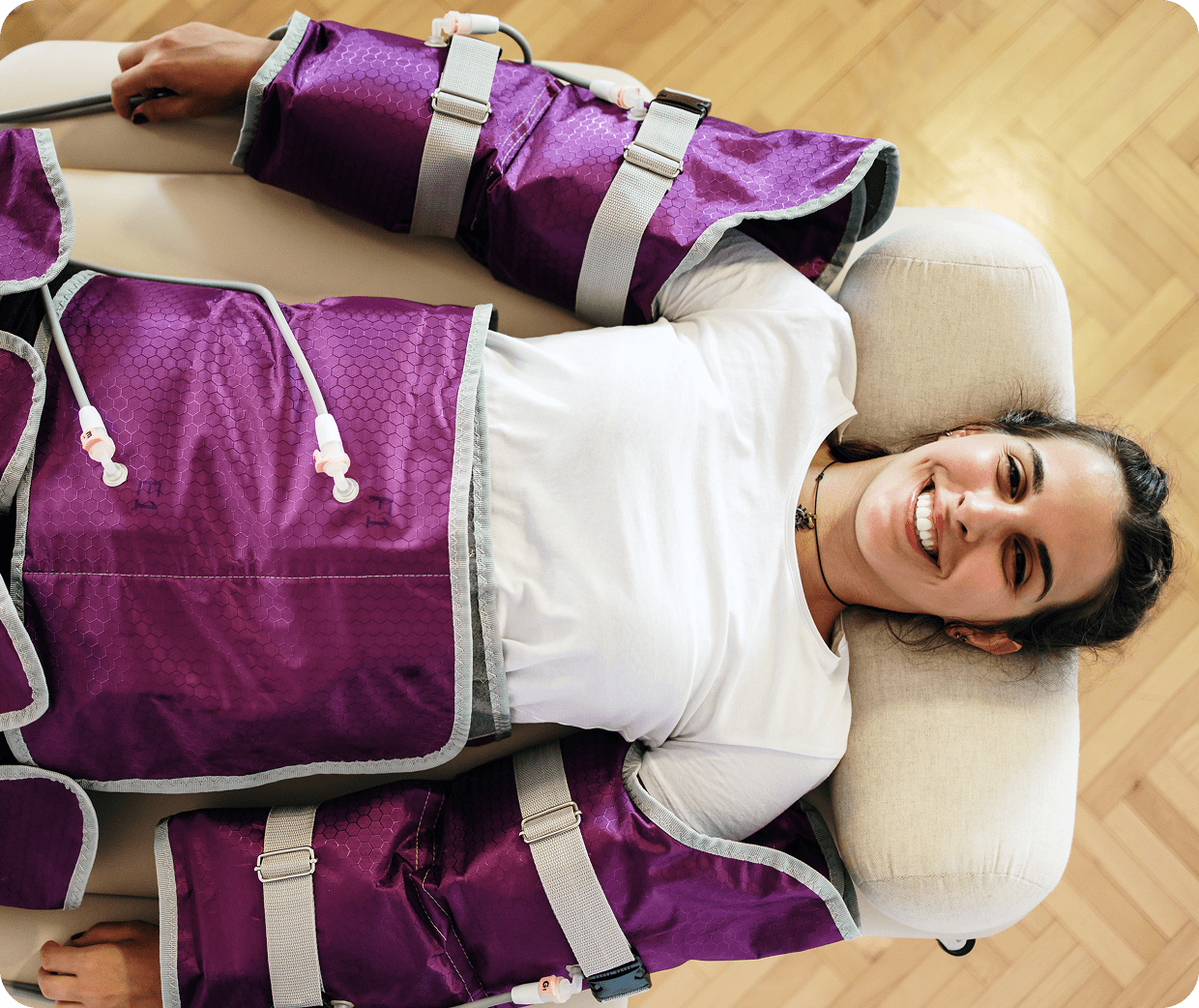 Person lying down, smiling, wearing purple compression sleeves on arms and legs, on a light-colored cushioned surface, in a room with a wooden floor.