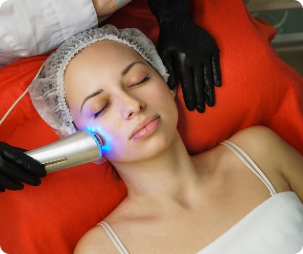 A woman with a hair cap and closed eyes receives facial treatment with a device emitting blue light, while lying on a red blanket. A person wearing black gloves holds the device.