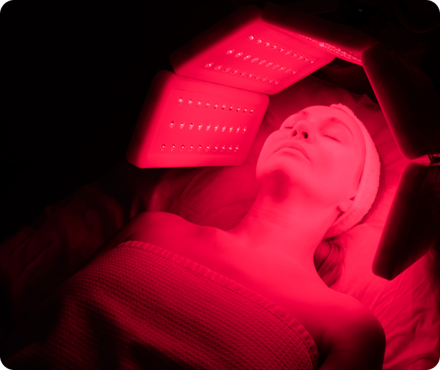 Person lying under red LED light therapy panels, wrapped in a towel, with eyes closed and headband on, in a dimly lit room.