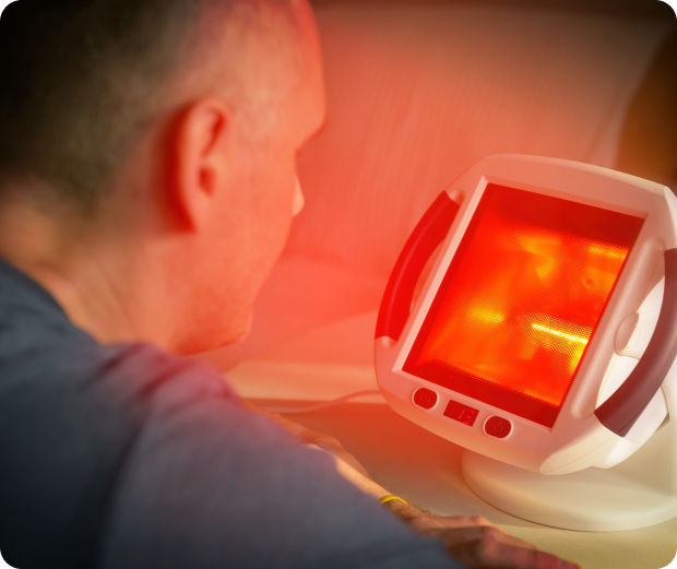 A man faces a glowing infrared heat lamp with a plastic casing, positioned on a table, emitting a red light.